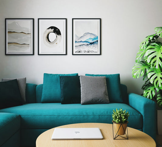 a dark teal couch next to an indoor plant under three pieces of wall art